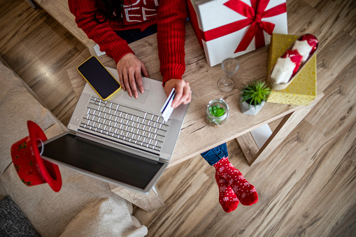 Woman use a credit card at home to buy New Year's gifts - A woman in a red sweater sits at a desk and uses a laptop and credit card to shop online at home