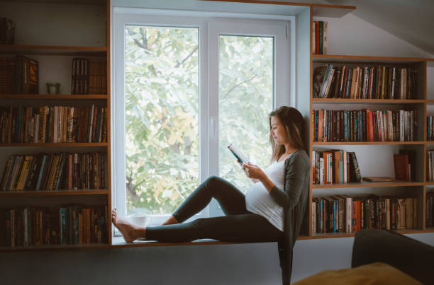 Pregnant woman enjoying on book reading. Full length pregnant woman reading books at home, she sitting around her domestic library. Books to Read During Pregnancy stock pictures, royalty-free photos & images