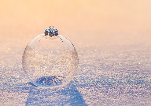 Christmas transparent bauble ornament covered with sparkling shiny snowflakes at sunset light on snow surface. Winter greeting cards natural blur blurred bokeh background