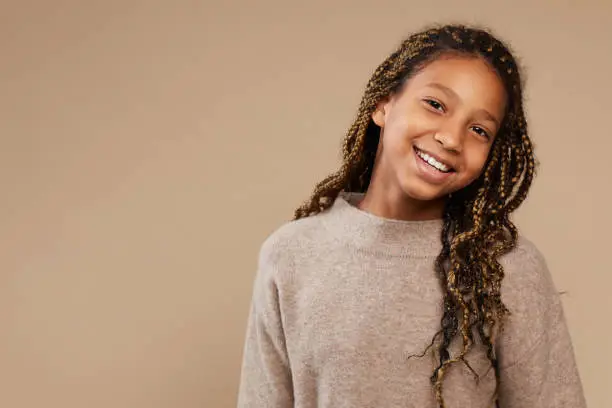Portrait of carefree African-American girl smiling happily at camera while standing against beige background in studio, copy space