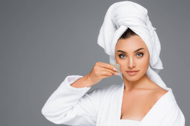 женщина с полотенцем на голове с помощью гуа-ша на лице изолированы на серый - spooning стоковые фото и изображения
