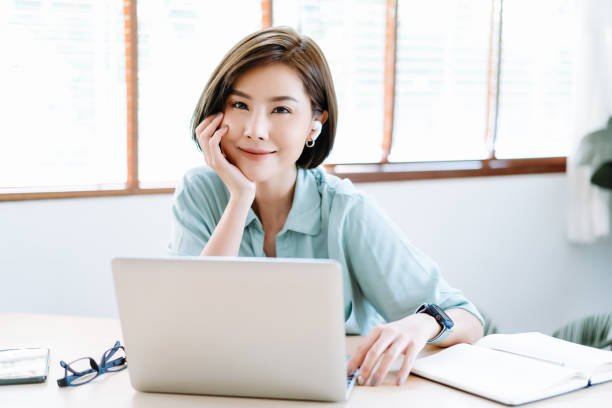 happy young beautiful asian freelancer woman using laptop  computer and wireless earbuds working remote at home.she communicating with service operator for online consultancy with customer. new normal - cotton swab audio imagens e fotografias de stock