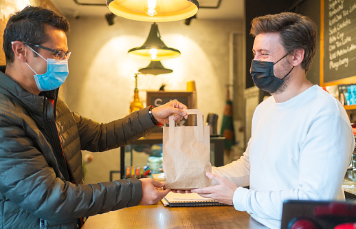 Happy male barista with protective face mask is giving orders in package to male smiling customer in modern cafe in new normal