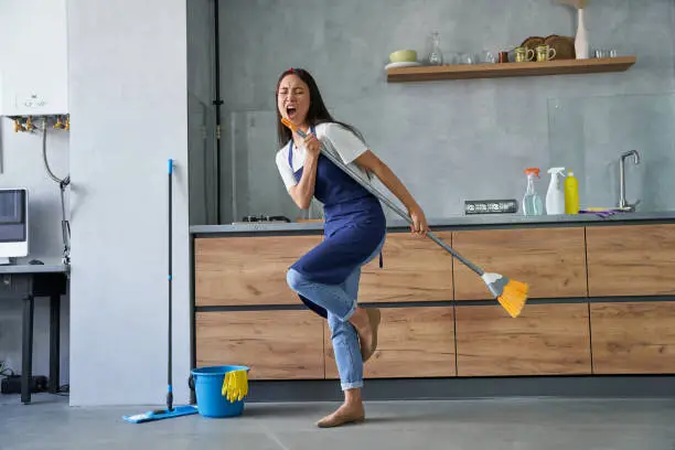 Full length shot of cheerful young woman, cleaning lady pretending to sing a song, holding broom while cleaning the floor, doing household chores. Housework and housekeeping concept