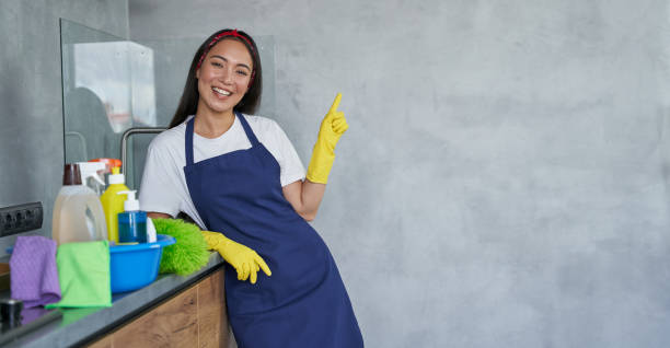 joven alegre, señora de la limpieza con guantes protectores sonriendo a la cámara, apuntando hacia arriba mientras está de pie en la cocina con productos de limpieza y equipo, listo para limpiar la casa - hygiene fotografías e imágenes de stock