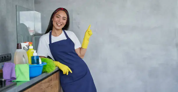 Cheerful young woman, cleaning lady in protective gloves smiling at camera, pointing up while standing in the kitchen with cleaning products and equipment, ready for cleaning the house. Housekeeping