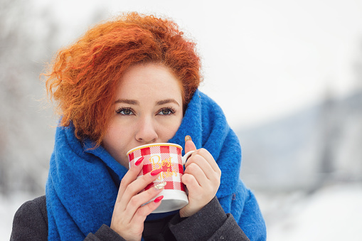 Young beautiful woman in warm clothing with cup of hot tea or coffee enjoying outdoors in the winter forest