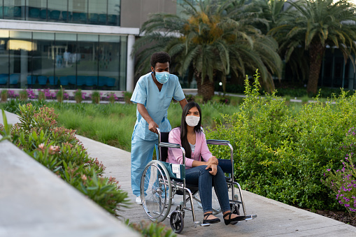 A black male nurse is carrying a young woman patient in a wheelchair through the hospital inner garden. New Normality.