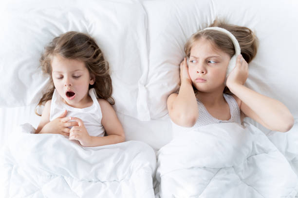 les petites filles dorment au lit. on ronfle dur, soeur branche des oreilles avec des écouteurs de fourrure. réveil tôt le matin, élévation à la maternelle, école. heure du coucher, rêves doux. les enfants corrigent la routine quotidienne pour l’ - pillow wake up yawning sleeping photos et images de collection