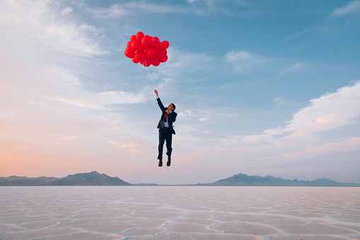 A businessman dressed in business suit flies away holding dozens of red balloons. He is looking to launch his business into the profits atmosphere. Image taken at the Bonneville Salt Flats in Utah, USA.