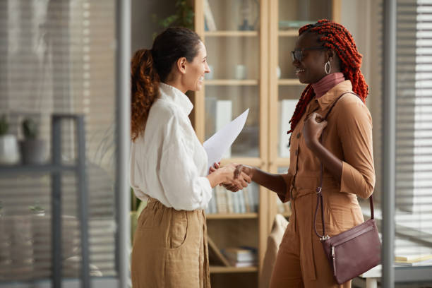 Two Young Women Shaking Hands at Job Interview Side view portrait of two successful young women shaking hands and smiling cheerfully while standing in modern office interior, copy space casual handshake stock pictures, royalty-free photos & images