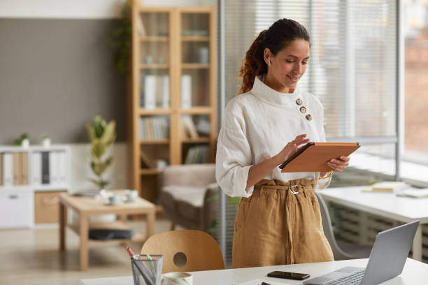 Modern Businesswoman Using Tablet Portrait of elegant businesswoman using digital tablet with wireless earphones while standing by desk in office or at home, copy space owner stock pictures, royalty-free photos & images