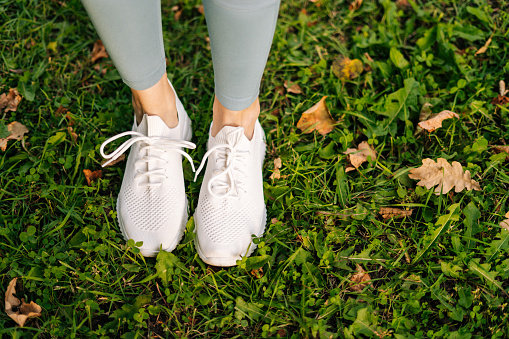 Close-up top view of unrecognizable woman feet in white sneakers standing and poses in place on green grass. Closeup view female feet in sneakers on green grass, closeup.