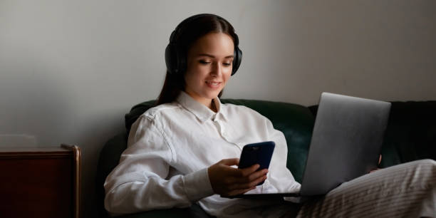 portrait of young woman who working on laptop computer indoors. unhappy girl using wireless headphones and mobile phone at home office. - credit crunch audio imagens e fotografias de stock