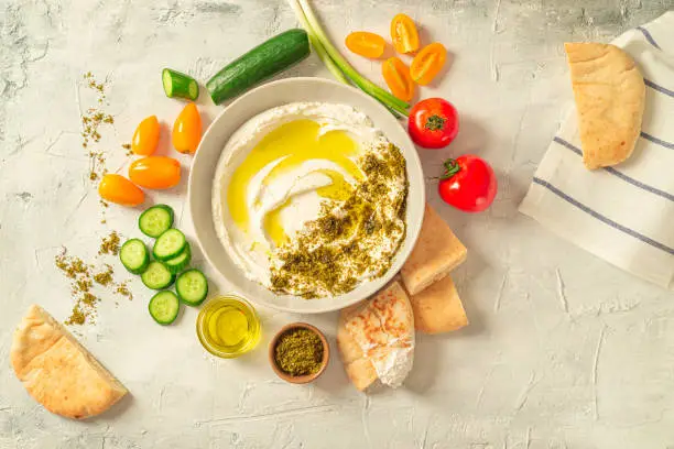 Photo of Popular middle eastern appetizer labneh or labaneh, soft white goat milk cheese with olive oil, hyssop or zaatar, served with many fresh vegetables on a wooden plate over grey table