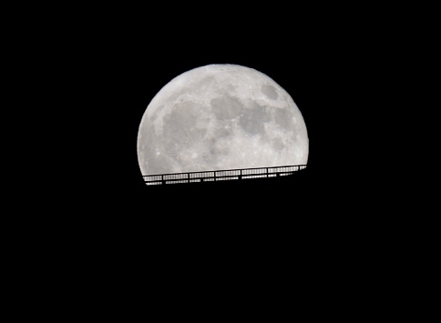This image shows a close up view of a magnificent full blue moon in the night sky above the silhouette of a roadway bridge.