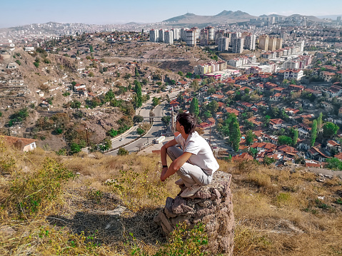 Young adult male tourist squatting on a stone looks at the panorama of Ankara (Turkey) from a height. Man admires the old slums and modern skyscrapers from the mountain in the Turkish capital center