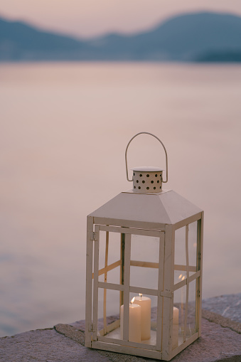 The body of a suspended metal lantern with three lighted candles on a stone border against a background of water and mountains. High quality photo
