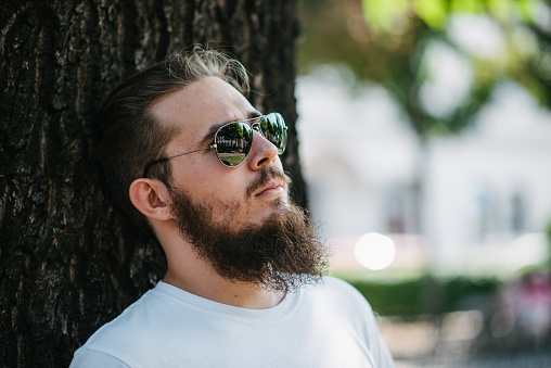 Portrait of a young hipster man leaning on a tree and relaxing.
