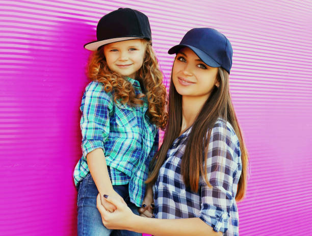 Portrait of young mother with child little girl wearing a checkered shirts and baseball cap in city over pink background stock photo