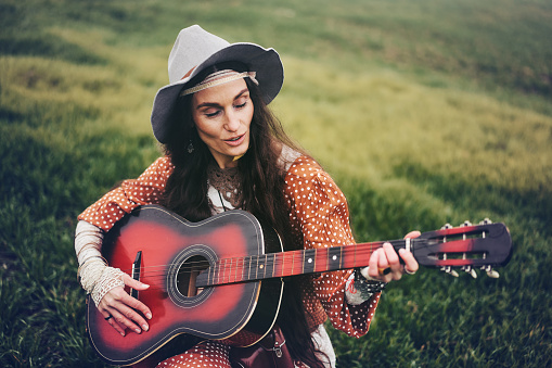 Mature hippie lady sitting on grass and plays guitar.