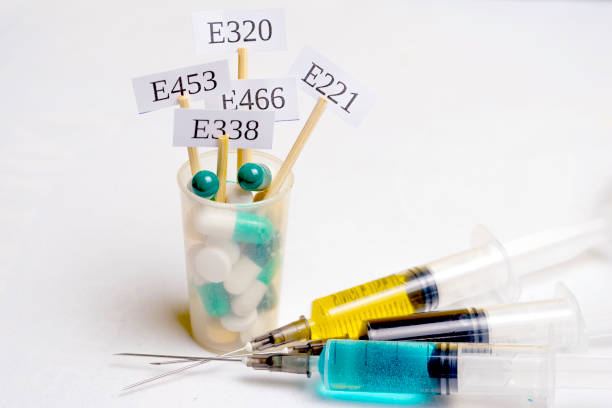 istock In a glass with multi-colored pills are plates with the code E-supplements. Nearby are syringes with nitrates, chemicals and additives. Harmful food additives. 1286105766