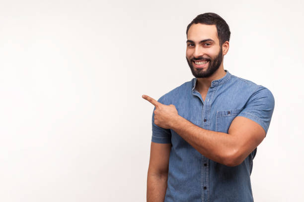 faites de la publicité ici! homme barbu positif pointant le doigt loin en prêtant votre attention à l’espace vide pour la publicité, regardant l’appareil-photo avec le sourire toothy - viser photos et images de collection