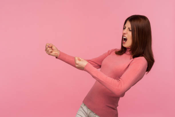 strong purposeful woman with brown hair in pink sweater making great efforts to achieve success, pretending to pull rope - imagination fantasy invisible women imagens e fotografias de stock