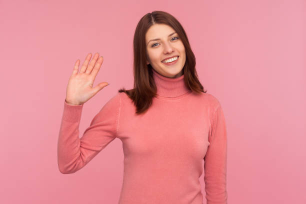 Friendly positive woman with brown hair waving her hand showing hi gesture, welcoming and greeting Friendly positive woman with brown hair waving her hand showing hi gesture, welcoming and greeting. Indoor studio shot isolated on pink background i 5 stock pictures, royalty-free photos & images