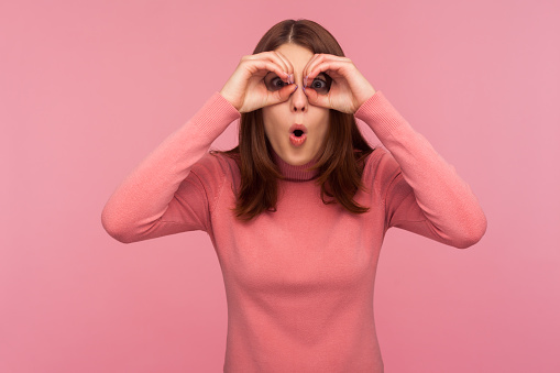 Funny curious woman with brown hair spying looking through holes in her hands, making binoculars, surprised with seen. Indoor studio shot isolated on pink background
