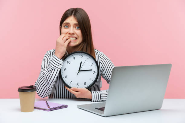 大きな壁時計を持って爪を噛む職場に座っているストライプシャツを着たせっかちな神経質な女性、締め切り - checking the time women impatient wristwatch ストックフォトと画像