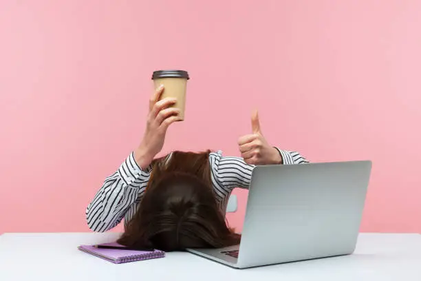 Photo of I am fine. Extremely tired brunette woman lying on desk at workplace holding paper cup of coffee and showing thumbs up, lack of energy, procrastination