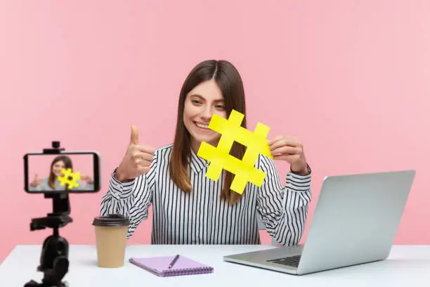 Photo of Positive smiling woman blogger recording video message on smartphone for her followers, showing yellow hashtag sign asking to rate post