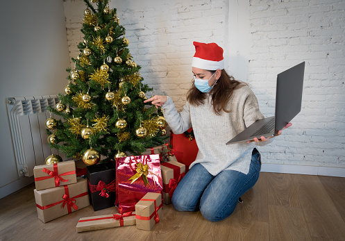 Happy woman with face mask home alone in lockdown celebrating christmas online video calling family and friends. Virtual holiday gathering and celebrations due to coronavirus second outbreak.