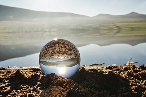 Pentland Hills, Edinburgh, Scotland - 6 November 2020: A glass ball has been used to create an inverted reflection of thin banks of mist drifting across Threipmuir reservoir on an otherwise sunny and still autumn day. The ball is resting on the sand of a small beach.