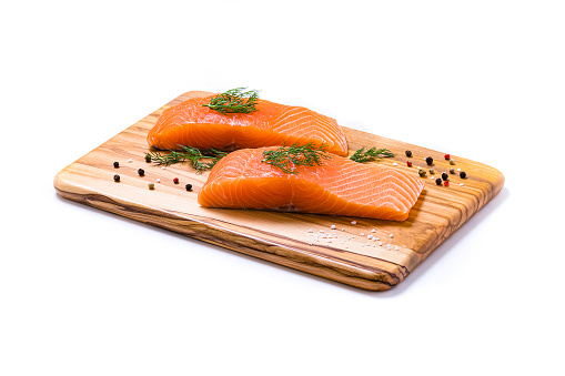 High angle view close-up of two salmon fillets on a wooden cutting board surrounded by some dill leaves and pepper. The cutting board is isolated on white background. Studio shot taken with Canon EOS 6D Mark II and Canon EF 100 mm f/ 2.8