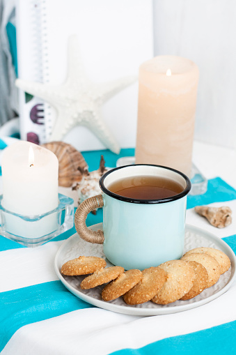 A study break: a hot mug of black tea with homemade cookies on a striped tablecloth; wax candles, decorative starfish, seashell, pile of notebooks on a white background.