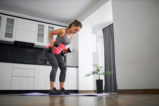 Side view of strong caucasian young woman training back on mat in kitchen. Fit girl wearing sportswear practicing at home in morning, lifting two dumbbells. Concept of home workout, weight loss.