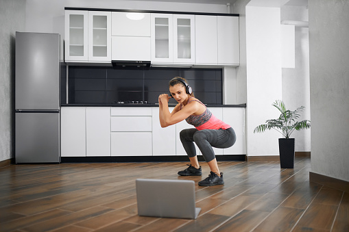 Side view of strong young woman squatting in kitchen, watching video on laptop. Fit girl wearing sports clothes and headphones training at home in morning, listening to music. Concept of home workout.