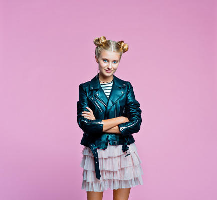 Confident female teenager wearing black leather jacket, pink tulle skirt standing with arms crossed. Studio shot on pink background.