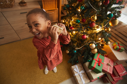 Fisheye portrait of excited African-American girl opening Christmas presents while standing by tree at home, copy space