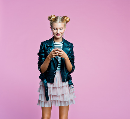 Female teenager wearing black leather jacket, pink tulle skirt and earphone using a smart phone. Studio shot on pink background.