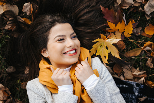 Portrait of a beautiful young woman with yellow scarf and irresistible smile lying in the fallen autumn leaves in park, top view