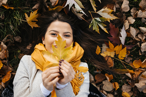 Portrait of a beautiful young woman with yellow scarf and irresistible smile lying in the fallen autumn leaves in park, top view