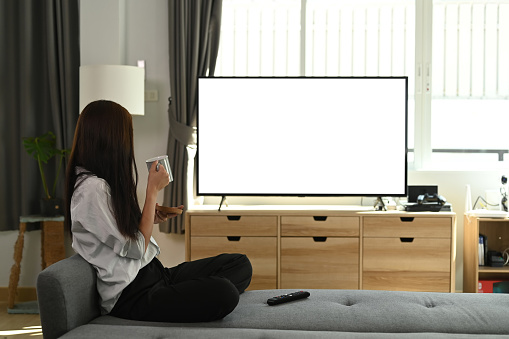 A young woman is drinking coffee and enjoying watching Television sitting on couch at home.