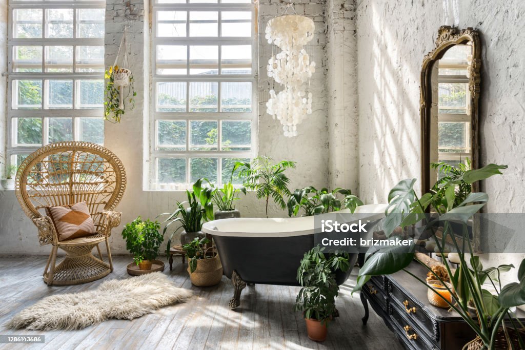 Cozy and comfortable room with interior in bohemian style Comfortable bathroom with interior design in boho chic style, bathtub, vintage commode with mirror, wicker armchair, fluffy carpet and green houseplants in flowerpots Bathroom Stock Photo