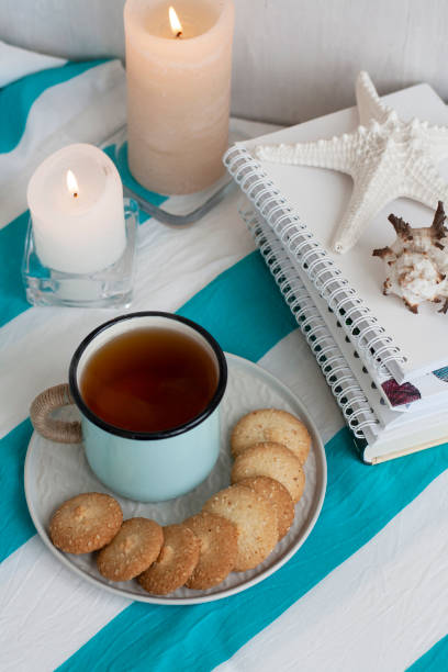 A hot mug of black tea with homemade cookies on a striped tablecloth, wax candles, decorative starfish, seashell, pile of notebooks on a white background.