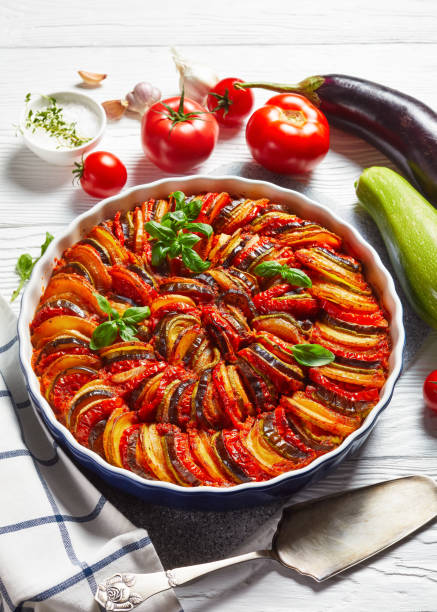 ratatouille, vegetable stew of sliced eggplant, zucchini, onion and potato with tomato sauce, ingredients at the background, french cuisine ratatouille, vegetable stew of sliced eggplant, zucchini, onion and potato with tomato sauce, ingredients at the background, french cuisine, vertical view from above ratatouille stock pictures, royalty-free photos & images