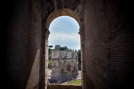 June 6, 2018 - Rome, Italy. Crowds of tourists walking around the Arch of Constantine as seen through the archways of the Roman Colosseum on a bright blue sky summer day.
