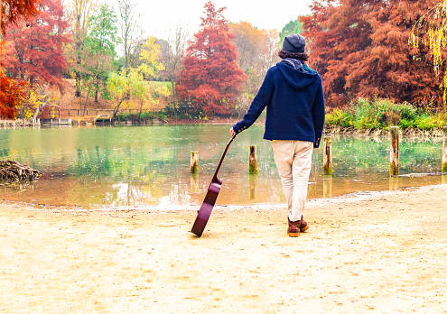 young curly latino man standing  alone in a park holding his guitar during autumn looking at a lake.red and orange mood.Beautiful landscape with yellow trees and Colorful foliage in the park.Falling leaves natural background.